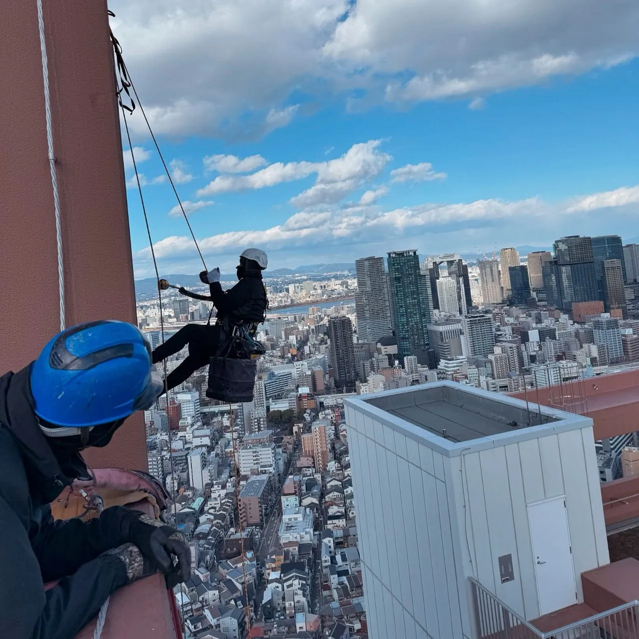 大阪の市街地を見下ろす高層ビルの屋上から、ロープで降下していく作業員と、屋上で支点を確保する作業員